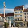 Das Hotel Zentral direkt am Hauptplatz, © Hotel Zentral Ein Platz mit einer Säule und Statuen, umgeben von Gebäuden, darunter das Hotel Zentral.