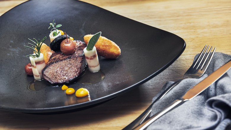 Pink glazed calf's liver, spelt balsam, Mangalitza bacon, Calvados apple, polenta dumplings, © Niederösterreich Werbung/Andreas Hofer Gourmet dish with veal liver, bacon, apple and polenta on a black plate.