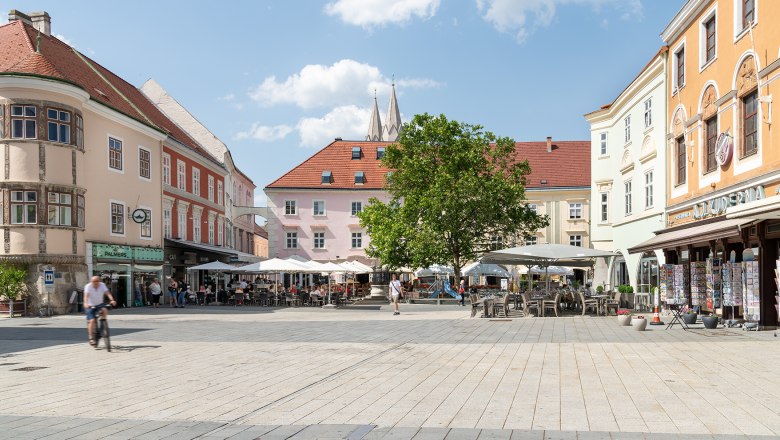 Hauptplatz mit Café Ferstl, © Wiener Alpen/Christoph Schubert Ein belebter Hauptplatz mit Cafés und Geschäften, umgeben von historischen Gebäuden. Menschen sitzen draußen, ein Radfahrer fährt vorbei. Blauer Himmel mit Wolken.