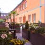 winrooms_gastgarten, © Winrroms Betriebs GmbH Ein sonniger Gastgarten mit orangen Gebäuden, Palmen und bunten Blumen in großen Pflanzkübeln.
