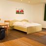 Studio, © Winrooms Betriebs GmbH Ein modernes Studio mit Doppelbett, Tisch und Stühlen, rotem Sessel und Koffer. Helle Wände, grüne Vorhänge und Blumenbilder an der Wand.