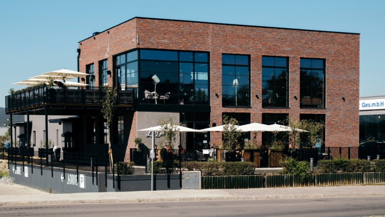 Le Burger, © Le Burger Modernes Backsteingebäude mit großen Fenstern und Terrasse mit Sonnenschirmen.