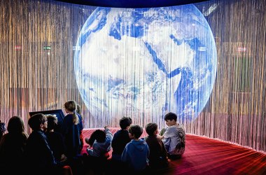 Kinder sitzen vor leuchtender Weltkugel-Projektion in einer Ausstellung.