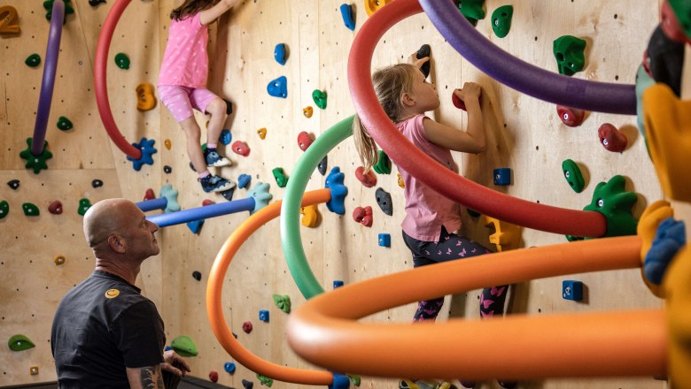 Kinder klettern an einer bunten Kletterwand mit Schaumstoffringen, während ein Erwachsener beaufsichtigt.