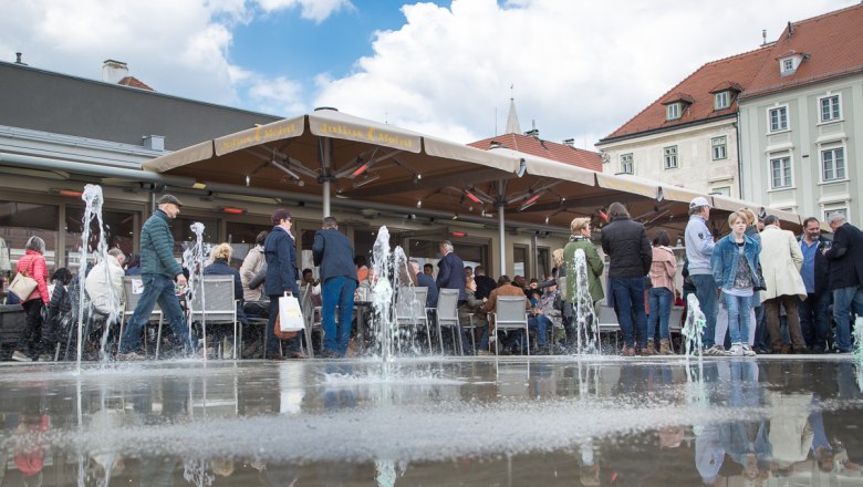 Menschen sitzen und stehen um einen Springbrunnen auf einem Platz mit Café.