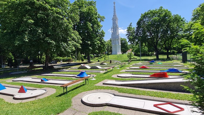 Minigolfplatz mit Hindernissen und einem hohen, weißen Turm im Hintergrund.
