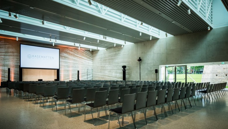 Moderner Veranstaltungsraum mit Stuhlreihen und großer Leinwand, beschriftet mit 'Kasematten Wiener Neustadt'.