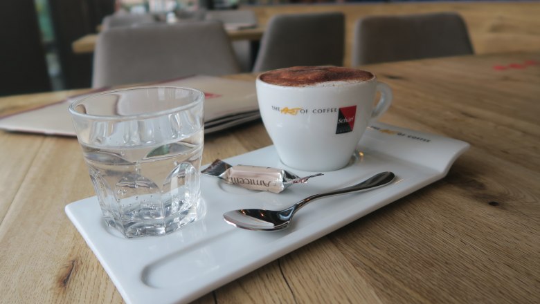 A cup of coffee with foam, a glass of water and a spoon on a tray in a café.