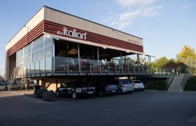 Modernes Restaurant mit Glasfassade und Terrasse, Autos parken darunter.