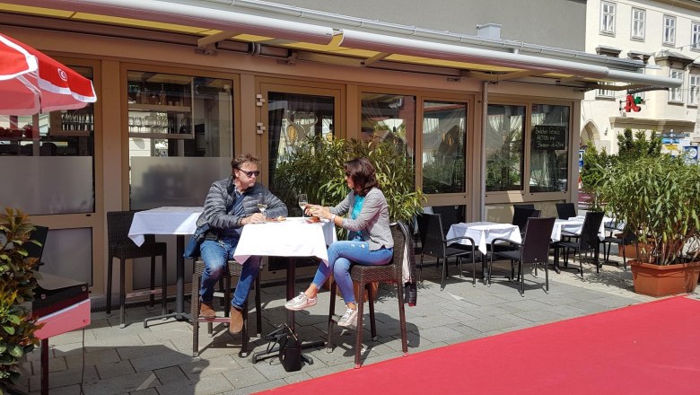 Two people are sitting at an outdoor table in front of a restaurant drinking wine.