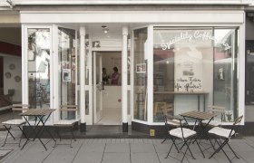 Außenansicht eines Cafés mit Tischen und Stühlen auf dem Gehweg.