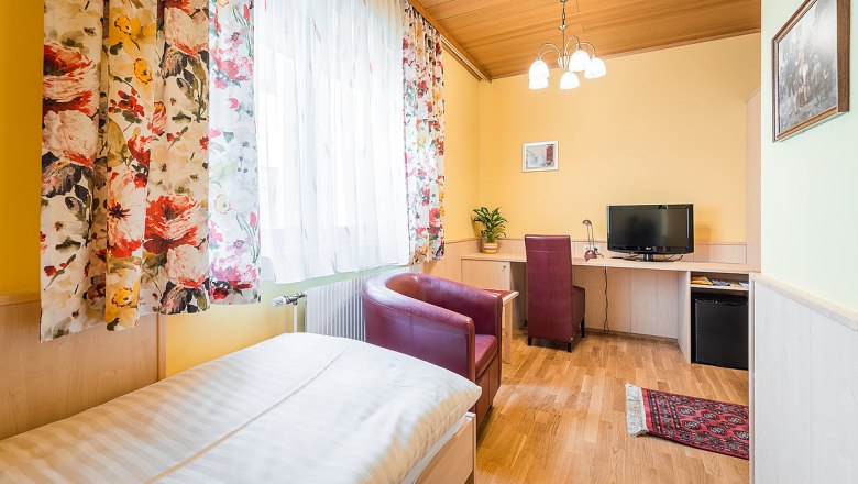 Ein gemütliches Einzelzimmer mit Holzboden, einem Bett, einem Schreibtisch mit Stuhl, einem Fernseher und bunten Vorhängen.