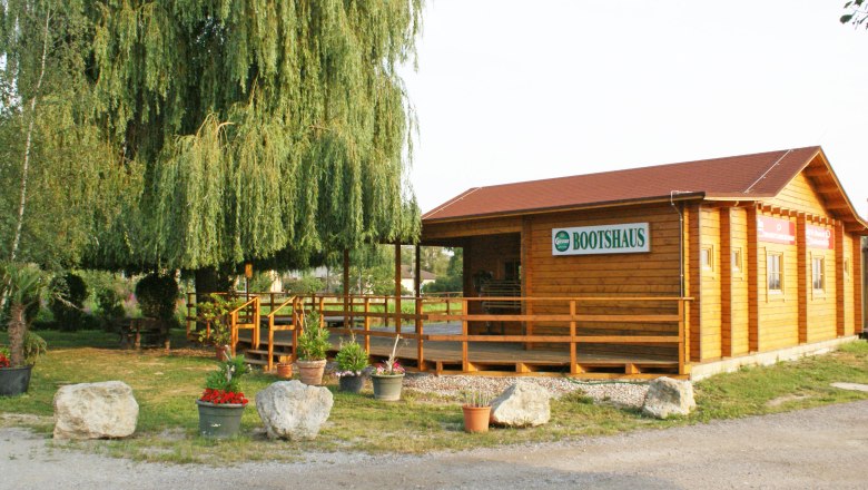 Holzgebäude mit Schild 'Bootshaus' neben einem großen Baum und Pflanzen.