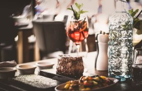 Ein saftiges Steak auf einem hei&szlig;en Stein serviert, begleitet von Gem&uuml;se, einer Karaffe Wasser und einem Cocktailglas.