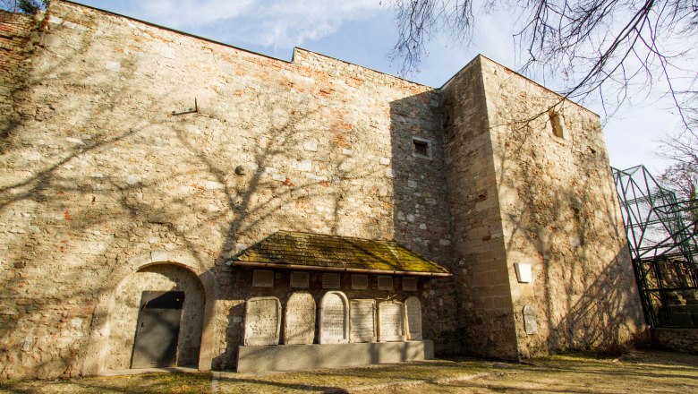 Steinmauer mit Torbogen und Gedenktafeln, Schatten von B&auml;umen auf der Mauer.