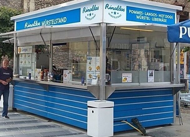 Ein blauer Imbissstand namens Rinaldas W&uuml;rstelstand mit Angeboten wie Pommes und Hot Dogs.