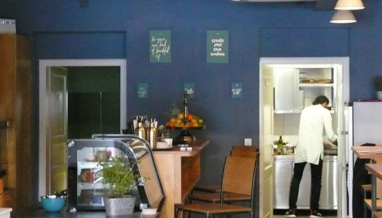 Innenansicht eines Caf&eacute;s mit blauer Wand, Theke, Barhockern und einer Person in der K&uuml;che.