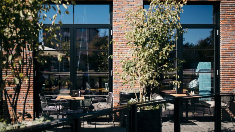 Ein gemütlicher Außenbereich eines Cafés mit Tischen und Stühlen vor einer Backsteinfassade, umgeben von Pflanzen.