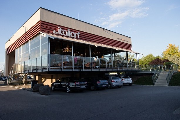 Modern restaurant with glass façade and terrace, cars park underneath.