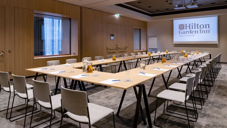 A modern seminar room with a U-shaped table arrangement, chairs, notepads and glasses. A screen with the Hilton Garden Inn logo on the wall.