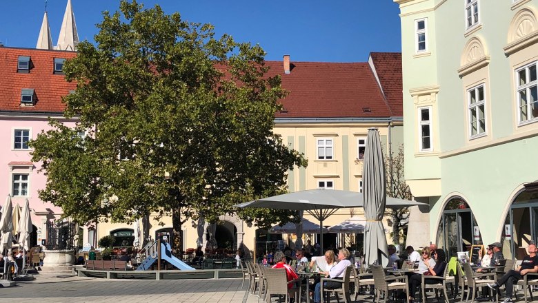Sonniger Hauptplatz mit einem Baum und Spielplatz beim Eterno. Menschen sitzen draußen und genießen das Wetter.
