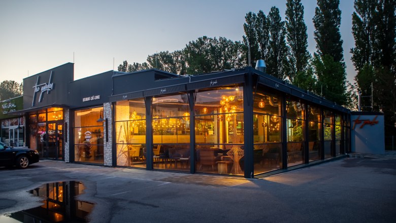 Außenansicht eines modernen Restaurants bei Dämmerung mit großen Glasfenstern und warmem Licht.