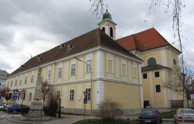 Historisches Gebäude mit gelber Fassade und rotem Dach, daneben eine Statue und parkende Autos.