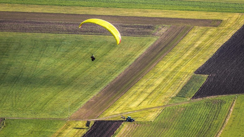 A parachutist with a yellow parachute over green fields, below a tractor on a dirt road.