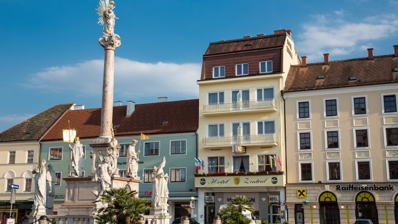 Ein Platz mit einer Säule und Statuen, umgeben von Gebäuden, darunter das Hotel Zentral.