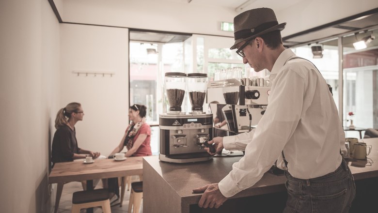 Ein Barista bereitet Kaffee in einem stilvollen Café zu, während zwei Frauen am Tisch sitzen und sich unterhalten.