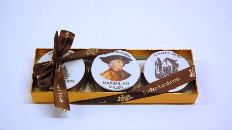 Eine goldene Schachtel mit drei Pralinen, verziert mit einem Bild von Maximilian und Wiener Neustadt Motiven, gebunden mit einem braunen Band.