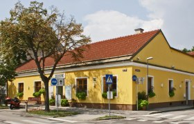 Gelbes Gebäude mit rotem Dach, beschildert als Gasthaus Weidinger, an einer Straßenecke mit Zebrastreifen und Verkehrsschildern.