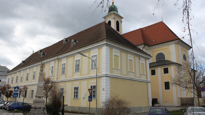 Historisches Gebäude mit gelber Fassade und rotem Dach, daneben eine Statue und parkende Autos.