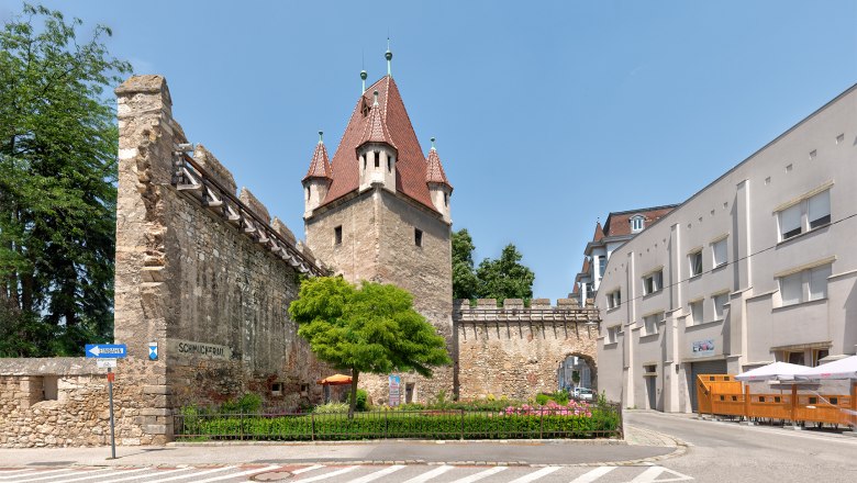 Historic stretching tower with red roof and adjoining wall, surrounded by modern buildings and a road.