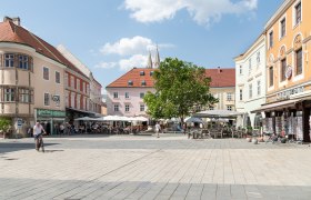 Ein belebter Hauptplatz mit Caf&eacute;s und Gesch&auml;ften, umgeben von historischen Geb&auml;uden. Menschen sitzen drau&szlig;en, ein Radfahrer f&auml;hrt vorbei. Blauer Himmel mit Wolken.