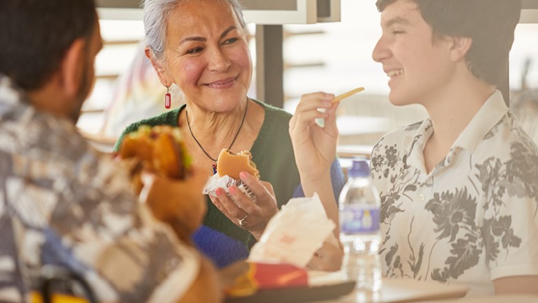 Drei Personen sitzen an einem Tisch und essen Fast Food. Eine ältere Frau lächelt einen jungen Mann an, der eine Pommes in der Hand hält.