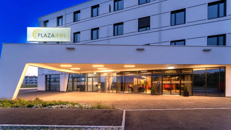 Eingang des Plaza Inn Hotels bei Nacht mit beleuchtetem Schild und moderner Architektur.