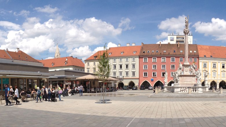 Panoramablick auf einen belebten Hauptplatz mit historischen Geb&auml;uden und einer Statue in der Mitte.