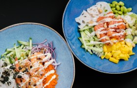 Zwei blaue Schalen mit hawaiianischen Bowls, gef&uuml;llt mit Lachs, Garnelen, Mango, Gurken, Edamame und So&szlig;en.