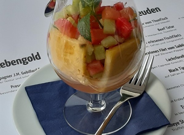 A glass of fruit salad on a plate with a blue napkin and cutlery.