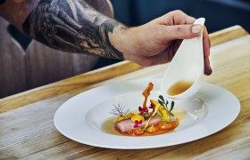 Ein Teller mit kunstvoll angerichtetem Essen, &uuml;ber das eine Hand aus einer kleinen Kanne Sauce gie&szlig;t.