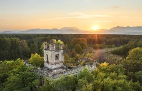 Verfallenes Gebäude im Grünen, im Hintergrund Sonnenuntergang über Bergen.