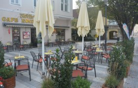 Ein leerer Schanigarten mit Tischen und St&uuml;hlen vor einem Caf&eacute; mit gelben Sonnenschirmen.