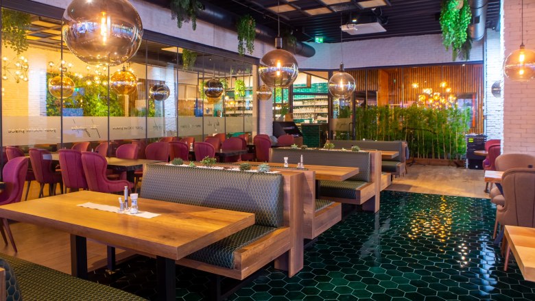 Innenansicht eines modernen Restaurants mit Holztischen, grünen Fliesen und hängenden Pflanzen.