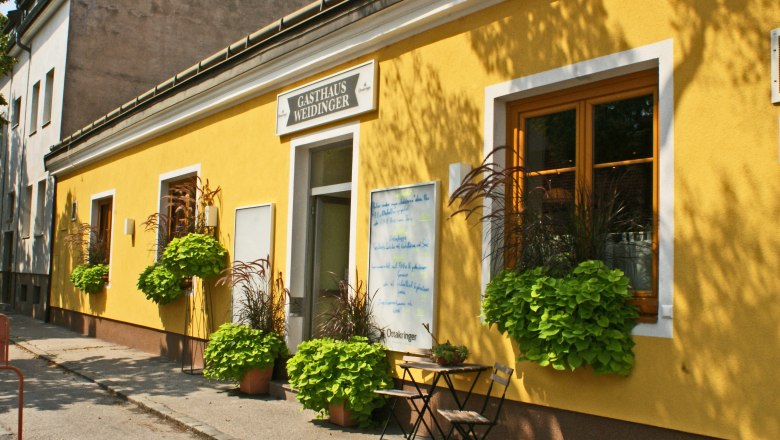 Gelbes Gebäude mit der Aufschrift 'Gasthaus Weidinger', Fenster mit Blumenkästen und ein Tisch mit Stuhl davor.