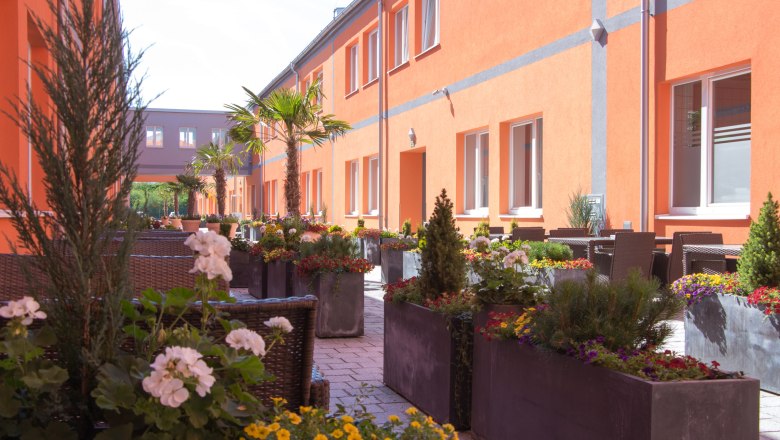 Ein sonniger Gastgarten mit orangen Gebäuden, Palmen und bunten Blumen in großen Pflanzkübeln.