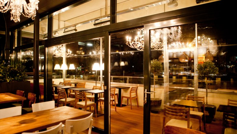 A stylishly furnished restaurant with wooden tables and chairs, illuminated by chandeliers and table lamps, overlooking a terrace.