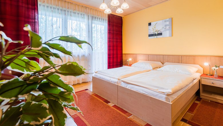 Gemütliches Schlafzimmer mit Doppelbett, gelber Wand, rotem Teppich und Pflanzen im Vordergrund.