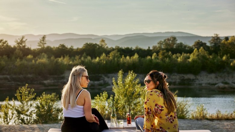 Zwei Frauen sitzen auf einer Decke am See, umgeben von Natur, bei Sonnenuntergang.