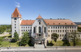 Historisches Gebäude mit rotem Dach und Turm, umgeben von Bäumen und einer Straße im Vordergrund.